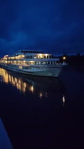 A large, multi-decked white ship named "KONSTANTIN KOROTKOV" is docked at a riverbank under a dark blue twilight sky. Lights are on in many of the ship's windows, casting reflections on the calm water. The ship is angled slightly towards the right side of the frame. The environment is dark, with the sky showing clouds and the water appearing black with illuminated reflections. The image is a still frame, likely from a video, capturing a moment of the ship at dusk or night.