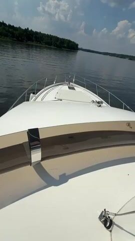 The video begins with a close-up view of the bow of a white yacht moving through calm, dark water. The camera angle is from the deck of the yacht, looking forward. Lush green trees line the distant shore. The sky above is a vibrant blue with fluffy white clouds. As the video progresses, the camera pans slightly, revealing more of the yacht's deck and the surrounding landscape. The boat continues to move smoothly across the water, creating gentle ripples. The scene is serene and peaceful, with the sounds of the boat engine and the water softly audible. The video concludes with a wider shot of the yacht sailing on the lake, with the shoreline still visible in the distance.
