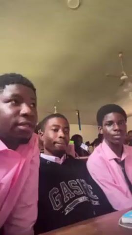 Three young men are seated in a classroom. The man on the left and the man on the right are wearing pink button-down shirts. The man in the middle is wearing a black hoodie. The man in the middle is holding a pen in his hand and pointing. The man on the left is filming the video. Other students are seated in the background. The classroom has a tan ceiling with a ceiling fan. The camera moves during the video.