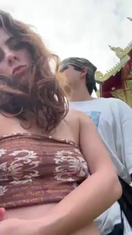 A young woman films herself with another person standing behind her and to her right. The woman is pale, with brown, wavy hair and wears a brown, strapless top printed with patterns and writing. She has a sullen expression. The person behind her is a young man with a mustache, wearing sunglasses and a camouflage bandana. He wears a white t-shirt with a graphic and a black cross-body bag. He is looking slightly away. They are outside, near a highly decorated Thai temple structure with gold trim. There is grass nearby. A dog is resting on the grass. The man shifts slightly and looks ahead. They are close to each other and walking slowly. The camera remains centered on the woman’s face, occasionally tilting slightly.