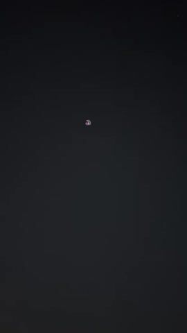 The video shows a dark sky with a few lights. In the first frame, a single red light is visible high up. In the second frame, two small white lights appear closer together and lower in the sky. Below the sky, a city skyline with buildings and lights is visible, with a blurry crowd of people in the foreground. It appears to be nighttime, possibly during an event, given the crowd and the lights in the sky.