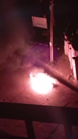 The video shows a nighttime scene from an elevated viewpoint, possibly a balcony or high window. A bright, circular red or orange flame is burning on the ground, producing a significant amount of white smoke. The surrounding area is dimly lit, with a few trees or bushes visible near what seems to be a street or path. A wooden post and a sign are also visible nearby. There are indistinct sounds that might be voices. The camera moves slightly, changing the perspective on the fire. The video ends without further change.