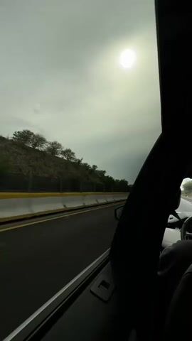The video shows a person driving a car on a highway. The sun is visible in the sky. The car is moving forward and the scenery outside the window is a blur. The camera then pans to the passenger, who is sitting in the passenger seat, wearing a hoodie. The passenger is looking at the camera and smiling. The video ends with a close-up of the passenger's face.
