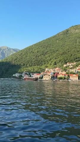 The video shows a wide view of a body of water in the foreground, with choppy waves reflecting the sunlight. In the background, a picturesque village is nestled against the base of a steep, densely forested green mountain. The village consists of multi-story buildings with terracotta roofs, lining the waterfront. A large, elongated boat with a dark hull and lighter superstructure is moored near the shore. The sky above is clear and blue, suggesting a sunny day. The camera appears to be moving, likely from a boat on the water, providing a panoramic perspective of the coastline.