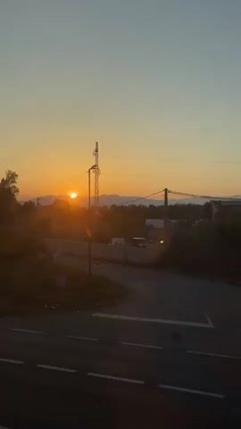 A sunset is visible with the sun low on the horizon. A telephone pole with wires and a street light are in the foreground. A car is driving on a road in the foreground. Trees and hills are visible in the background.