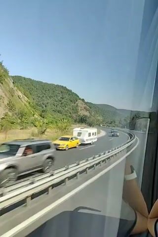 The video shows a view from inside a vehicle, looking out a window at a highway. A white recreational vehicle is on the left side of the road, driving in the same direction as the viewer's vehicle. Further down the highway, a dark-colored car is visible. The highway curves to the right, with a steep, tree-covered hillside on the left. The sky is clear and blue. A person's arm and wrist are visible in the lower right corner of the frame, suggesting the footage is from a passenger in a vehicle.