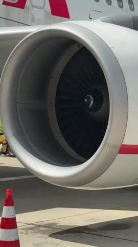 A close-up shot shows an airplane engine. The engine is a turbofan engine, with a large fan at the front. The fan blades are visible and appear to be made of metal. The engine is white with red accents, and the airline's logo is visible on the side of the plane. The engine is mounted on the wing of the airplane. In the background, a traffic cone with red and white stripes is visible on the tarmac. The sky is cloudy. The video is about four seconds long and shows the engine from a slight angle.