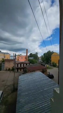 The video shows a street scene in a small town or city. In the foreground is a corrugated metal roof. Beyond it, an empty parking lot is visible, with a silver minivan parked in it. A utility pole stands in the center of the frame, with wires running from it. There are two pigeons on the wires, one on the lower wire and one on the higher wire atop the pole. In the background are several buildings of varying sizes and colors, including yellow, orange, and brick. Two brick smokestacks rise above the buildings. The sky is cloudy, with patches of blue visible.