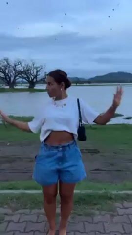 A woman stands near a lake on a cloudy day, dressed in a white T-shirt, blue denim shorts, and bare feet. She has her arms outstretched, palms up. There are trees and mountains visible in the background. A flock of birds flies overhead. The woman is wearing a necklace and carrying a small black bag over her shoulder. She looks to her right. She is standing on a paved path next to a grassy area.