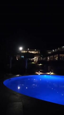 Here's a description of the video:
The video shows a swimming pool, illuminated with blue lights, at night. In the background, there is a building with multiple levels. There are also small lights around the pool area. The sky is dark with what could be stars visible. The scene appears peaceful and quiet.