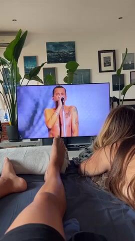 This video shows a close-up view of two people on a bed watching a television. The person in the foreground is lying on their back, with their legs extended and feet visible. Their feet are bare and one leg is propped up on a white pillow. The second person is lying on their side, facing away from the camera, with their head resting on their arm. Their long, blonde hair is visible. The television screen displays a man singing into a microphone, with bright pink and blue stage lighting. The room appears to be a bedroom, with a large potted plant on the left and several framed pictures on the wall behind the television. The overall lighting is dim, with the primary illumination coming from the television screen.