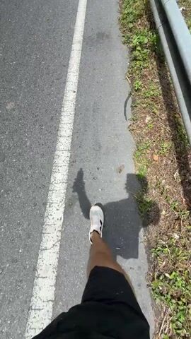 The video shows a person walking on a paved road alongside a white painted line, with a metal guardrail to the right and lush greenery lining the side. The road leads downhill toward a body of water visible in the distance. Overhead power lines run along the roadside, supported by utility poles. The sky is visible and partially obscured by clouds. A white van drives toward the camera.