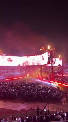 Here's a description of the video:

The video shows a large stadium concert at night. The stage has multiple large screens displaying the performers. There are pyrotechnics going off in the form of bursts of flames above the stage and along the front of the stage. The audience is huge, and most of them are holding up what appear to be light sticks. The lighting is predominantly red.
