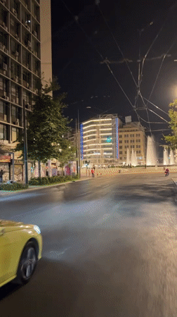 Wet Athens streets buzz with taxis, traffic, and a lit fountain
