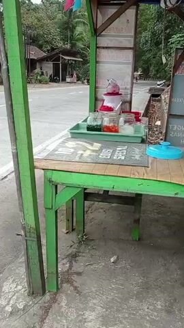 A food stall is set up on the side of a road. The stall is painted green and has a wooden table covered with a mat and various food items. There are jars of colorful liquids, possibly syrups, and a red dispenser. A blue lid is also visible on the table. Behind the stall, there is a road with a few buildings and trees visible in the background. The ground is dirt and gravel. The video appears to be taken from a stationary position, focusing on the stall.