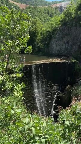 A waterfall cascades down a layered rock face. Above the waterfall, a body of water is visible, and on its edge, two figures stand. One figure, wearing a blue swimsuit, is bent over, while the other, in a white swimsuit, is standing upright. The area surrounding the waterfall is lush with green foliage and trees, with a rocky cliff face behind the water. The sky above is clear and blue.
