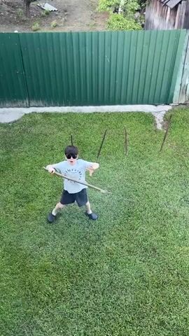Two young boys are playing in a grassy backyard. On the left, a boy in a white t-shirt, dark shorts, and dark sandals is near a basketball. On the right, another boy in a light-colored t-shirt, dark shorts, dark sandals, and sunglasses holds a long stick horizontally across his body, positioned in front of four shorter sticks vertically planted in the grass. The boy with the basketball moves it with his feet towards the planted sticks, while the boy with the long stick pivots and sweeps the stick downwards to block the ball's path. A green corrugated metal fence is visible in the background.