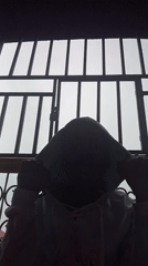 Hooded figure behind barred window, Nkpor rooftops in background