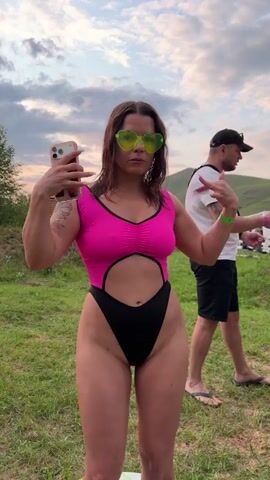 A woman in a pink and black one-piece swimsuit and heart-shaped sunglasses poses for a photo while holding a smartphone. She is standing in a grassy outdoor area. In the background, a man in a white t-shirt and black shorts walks away from the camera. The sky is cloudy, suggesting it might be late afternoon or evening. The setting appears to be a festival or outdoor gathering, with tents visible in the distance. The woman has a confident and playful expression.