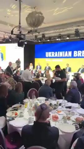 The scene is a formal indoor event with a seated lunch or dinner. A man in a white shirt serves food from a tray to seated individuals around a table. Behind the tables, people sit on a raised platform, some with legs crossed, against a backdrop with the words "Ukrainian Davos, Switzerland" on a yellow and blue background. The atmosphere appears to be a business or political gathering. The room is brightly lit with a chandelier overhead and camera equipment is visible.
