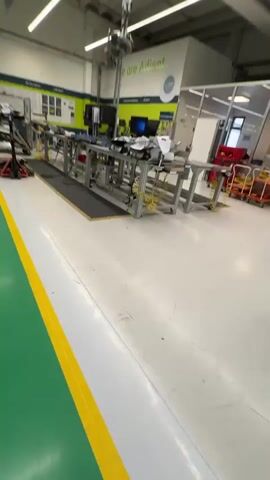 The video pans across a brightly lit industrial workshop. The floor is a clean white, with green and yellow safety lines demarcating pathways. To the left, a wall features a large screen displaying graphics and text. Further into the room, a long workbench is set up with various electronic components, tools, and equipment, including computers and machinery. A mechanical arm is suspended from the ceiling above the workbench. In the foreground, a pallet jack is being used to move a stack of car seats, demonstrating an ongoing assembly or logistics process within the facility. The overall environment suggests a modern, organized manufacturing or testing space.