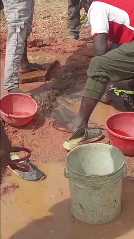 The video captures several men engaged in an outdoor activity within a muddy, reddish-brown environment in Agingi Zion, Nigeria. The scene is centered around a shallow, dug-out pit or trench filled with muddy water and sediment.

Four men are prominently visible. One man, wearing a red, white, and black striped shirt over dark green athletic pants (with a "Hummel" logo), is kneeling and reaching deeply into the muddy water with both hands, seemingly sifting or searching through the sediment. Another man, dressed in a black hoodie featuring a "NIKE" logo and light grey cargo pants, stands barefoot near the pit, his feet partially submerged in the muddy water. A third man, seen from behind in a white tank top and dark shorts, stands by the edge of the pit. Further in the background, another man in a brown jacket crouches, also interacting with the muddy area.

Several objects are scattered around the work area: two red plastic bowls, one appearing to hold muddy water and possibly some small items, and a larger, dirty, light-colored plastic bucket. Two pairs of sandals or slides (one dark grey, one light yellowish-green) lie partially submerged in the muddy water, suggesting they were removed before the men entered the pit.

The ground is a distinctive reddish-brown, indicative of lateritic soil, and wet from the water. In the background, sparse green vegetation is visible, alongside distant buildings and rocky hills, under a bright, sunny sky. The presence of clear shadows indicates daytime. The men appear focused and engaged in their manual task, which seems to involve careful sifting or washing of material from the muddy water. No strong emotions are overtly displayed, only concentration on the activity.