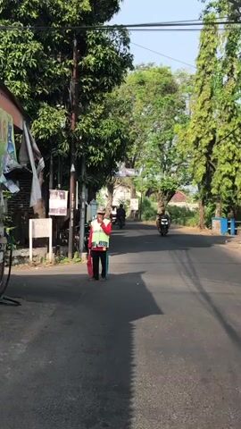 A man wearing a red and white vest, a white hat, and dark pants stands on the side of a paved road. He is holding a red bag and a white object. The road is lined with trees on both sides. In the background, two motorcycles are visible, and a person is riding each motorcycle. The lighting suggests it is daytime, likely morning or afternoon, with shadows cast on the road. There are some structures and posters on the left side of the road. The overall environment appears to be a rural or suburban street.