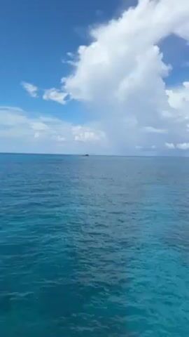 The scene shows a vast expanse of turquoise ocean under a clear blue sky dotted with fluffy white clouds. In the distance, a small, dark shape possibly a boat or island is visible on the horizon. The water is slightly choppy, and its color varies from light turquoise near the surface to deeper shades of blue in the distance.