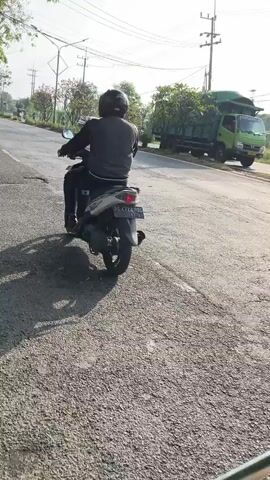A person wearing a helmet and a dark jacket rides a silver scooter on a paved road. A green dump truck is visible in the background on the right side of the road. The scene appears to be outdoors during the day, with sunlight casting shadows.