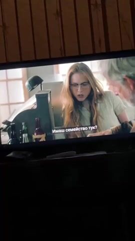 The video shows a television screen displaying a scene from a movie or TV show. On the left, a woman with long, light reddish-brown hair and glasses, wearing a light-colored, possibly checkered shirt, is focused downwards, her expression serious. To her right, a man with gray hair and a beard, dressed in a plaid shirt, is pouring liquid from a bottle into a glass. The two are positioned at a counter, which holds several bottles, a dark helmet, and what appears to be an old cash register or piece of equipment. The setting within the show appears to be an indoor establishment, possibly a shop or diner, with light walls and windows in the background. Bulgarian subtitles are visible on the screen, translating dialogue as: "Имаш семейство тук?" (Do you have family here?), "Е, сега е само синът ми." (Well, now it's just my son.), and "Той дойде тук с жена ми и дъщеря ми..." (He came here with my wife and my daughter...). The television is in a room with striped wallpaper behind it.