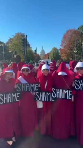 The video shows a group of people protesting in the street, likely in Washington D.C., as the US Capitol building is visible in the background. The protesters are dressed in red robes and white bonnets, resembling the characters from "The Handmaid's Tale." They are holding signs that read "Shame" and chanting the same word. The sky is blue, and the weather appears clear and sunny. The street is paved and lined with trees, some of which have autumn colors.