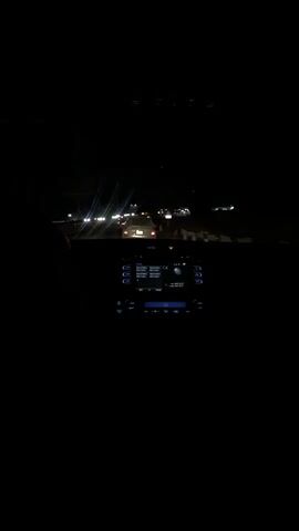A passenger's perspective shows a car driving at night. Visible in the windshield are tail lights of other vehicles and city lights in the distance. The dashboard and console are visible in the foreground. Music is playing.