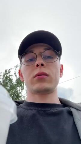 A close-up shot of a young man wearing a black baseball cap and clear-rimmed glasses, looking upwards and to the right. He is wearing a black t-shirt and a quilted gray jacket. His left hand is visible in the foreground, holding a white plastic cup. The background is a blurry, overcast sky with some green foliage visible to the left. The man's face is in sharp focus, and his lips are slightly parted. The video appears to be filmed from a low angle, looking up at the man.