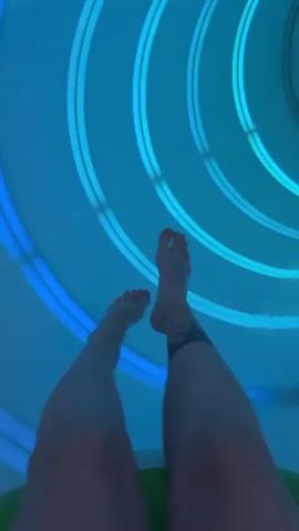 A first-person view shows a pair of legs descending a water slide while in a flotation device. The walls of the slide are white with darker outlines. Light fixtures shine blue light inside the slide. The bare legs move through the slide at a fast pace. One of the legs has a tattoo around the ankle. The person is yelling in excitement.
