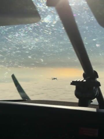A small airplane flies over a layer of clouds. The sun is setting or rising, casting a warm glow in the sky. The camera is inside another aircraft, looking out of the cockpit window. A propeller or part of the aircraft is visible in the foreground.