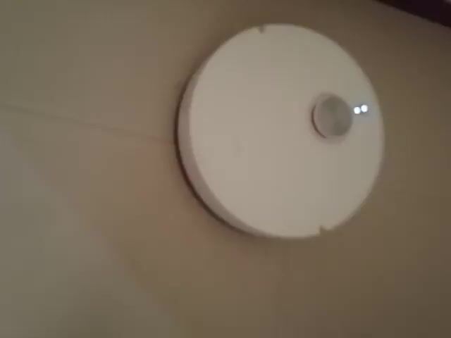 A white robotic vacuum cleaner with two indicator lights on top moves across a tiled floor. The vacuum cleaner is round and has a silver dial in the center of its top surface. The floor is beige and appears to be made of large tiles. The lighting is dim, and the image quality is poor. The vacuum cleaner moves from left to right in the frame.