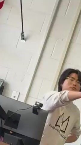 A young man with dark hair and glasses stands in what appears to be a classroom. He is wearing a white hoodie with a black design on the front. He is gesturing with his right hand while looking towards something or someone outside the frame. A black computer monitor and a webcam are visible on a desk next to him. The wall behind him is white and constructed of rectangular blocks. There is a metal rod mounted on the wall near the top of the frame.