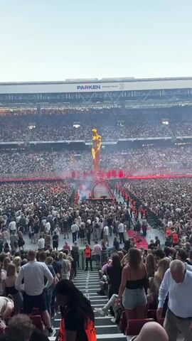 A wide-angle shot captures a large outdoor stadium filled with spectators. In the center of the field, a massive, golden, humanoid statue stands tall. Surrounding the base of the statue is a circular stage with red lighting and golden accents. Several performers dressed in flowing red robes are positioned on or around the stage, some appearing to be walking towards or away from the statue. Smoke or fog effects are visible around the stage. The crowd, densely packed, fills the stadium seating and the area around the stage, with many people holding up their phones, likely recording the event. The stadium appears to be a sports venue with tiered seating and large screens visible in the background. The lighting suggests it is late afternoon or early evening.