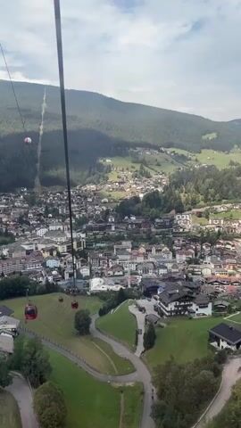 A first-person perspective video shows a view from a cable car ascending a mountain. Below, a picturesque town is nestled in a valley, surrounded by lush green hills and dense forests. Numerous red cable cars are visible, some in the foreground and others further away, following the aerial route. The town features a mix of traditional buildings and more modern structures, with a church spire prominently visible. Roads wind through the town and up the hillsides, and some patches of farmland are scattered throughout the landscape. The sky is partly cloudy. The audio contains speech in Mandarin, with the speaker mentioning waking up at 8:30 AM for an 8:30 AM appointment.
