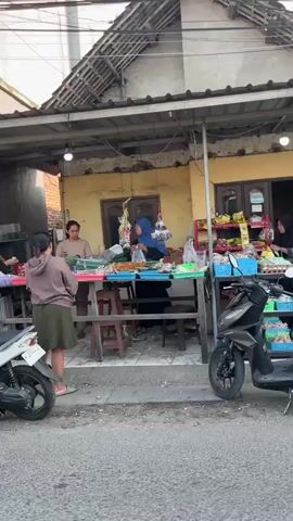 The video shows a street-level view of a small food stall with a covered outdoor seating area. There are three women and one person whose gender is not clearly discernible visible at the stall. Two women in hijabs are on the left, and two people are standing behind a counter on the right, where food items are displayed. A white scooter is parked in front of the stall, and an orange scooter is parked to the left. The stall appears to be made of wood and is attached to a building with a yellow facade. The overall environment suggests a casual, outdoor marketplace or roadside vendor setting.