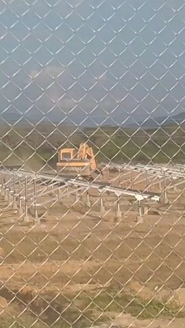 The video shows a yellow Hyundai excavator moving dirt on a construction site, partially obscured by a chain link fence. The site features a grid of metal supports. The environment appears to be a dry, grassy field with a line of trees in the background. The sky is overcast.