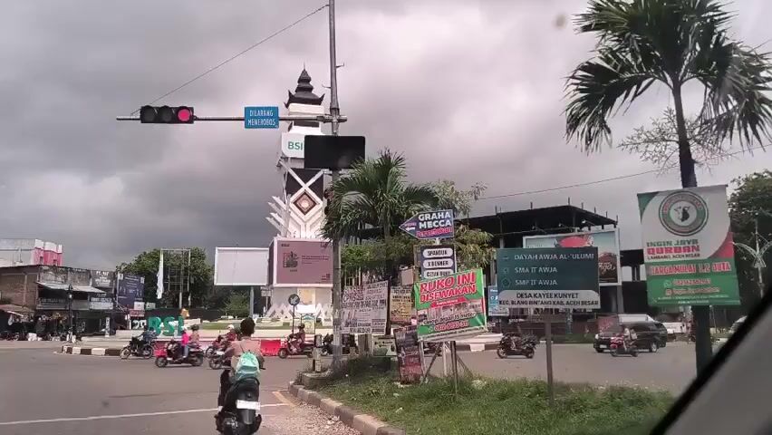 The video shows a street intersection in a town. The traffic light is red, and several motorcycles are waiting at the intersection. A white car is stopped at the intersection as well. There is a monument in the center of the intersection, surrounded by billboards and signs. The sky is overcast and cloudy. Buildings line the street in the background.