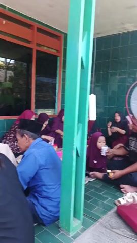 In the first scene, a group of people are gathered around a table in what appears to be a covered outdoor area. Some are seated on the floor, while others are on chairs. One person, a woman wearing a hijab, is feeding a young child who is also wearing a hijab. There is a table with drinks and snacks. The walls are tiled with green tiles. A fan is visible in the background. In the second scene, the same group of people are gathered, but the setting appears to be slightly different, possibly the same area but with a different arrangement. A woman in a light blue hijab is standing and appears to be speaking animatedly. A child is seated in front of her, and other people are seated around. In the third scene, a woman in a light pink dress is handing a cup to another woman wearing a blue hijab. Other people are seated on the floor. In the fourth scene, the woman in the pink dress is still serving drinks, with other women seated. In the fifth scene, a man in a patterned shirt and black pants is standing and looking away. Another woman in a hijab is standing in the background, near a table with drinks. Shoes are scattered on the floor. The overall scene suggests a social gathering, possibly a family event or a religious ceremony.