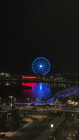 The video shows a nighttime aerial view of a city with a large Ferris wheel illuminated by colorful lights. The Ferris wheel is the central focus, with its blue and then green lights contrasting against the dark sky. Below the Ferris wheel, there is a body of water with reflections of the lights, creating a vibrant red and blue effect. In the foreground, there are city streets with visible traffic lights and palm trees. A modern, arched bridge is also illuminated with blue lights. The overall scene depicts a lively urban landscape at night, highlighting the illuminated Ferris wheel and its surroundings.