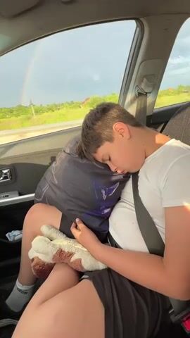 A young boy is sleeping in a car. He is wearing a white t-shirt and black shorts. He is holding a stuffed animal. A pillow is behind his head. A seatbelt is across his chest. Outside the car, a rainbow is visible in the sky. The car appears to be in motion.