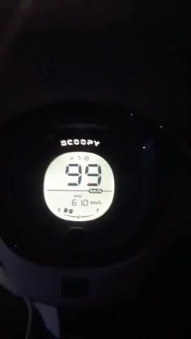 The video shows a dashboard of a motorcycle at night. The dashboard displays the brand name SCOOPY and the speed of the motorcycle, which increases from 100 km/h to 103 km/h. The video also shows the fuel gauge and the average fuel consumption. The background is dark, indicating it is night. The road ahead is visible with streetlights in the distance.