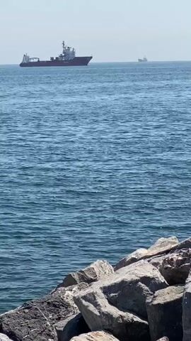 The video shows a wide shot of the sea with several ships moving across the frame. The sea is blue with small ripples on the surface. In the foreground, there are large rocks, some wet from the water. In the distance, a large red and white ship moves from left to right, followed by a darker ship moving from right to left. Another dark ship is visible in the far distance, moving from left to right. The sky is bright and clear.