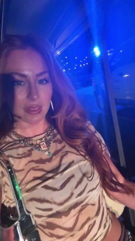 A woman with reddish-brown hair, wearing a tiger-striped sheer top and layered necklaces, sits inside a golf cart-like vehicle. The lighting is dim with a blue hue. Other people are visible in the back seats of the vehicle. The woman smiles and speaks to the camera as the vehicle moves. A shiny purse hangs from her shoulder.  Streetlights are visible through the windows, suggesting it is night. The vehicle appears to be a shuttle of some kind.