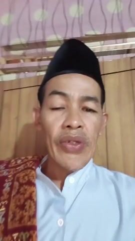 A man wearing a black cap and a light blue shirt with a red and gold patterned cloth draped over his left shoulder speaks to the camera. He has a mustache and appears to be winking with his right eye. The background consists of a patterned wallpaper and wooden paneling. The timestamp at the beginning of the video is 00:00.