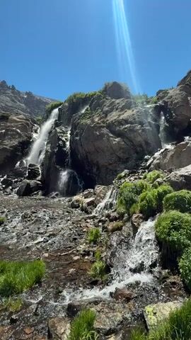 A person with a backpack and a white cap is walking on a rocky path towards a waterfall. The waterfall is surrounded by rocks and vegetation. The sun is shining brightly in a clear blue sky. There is also some snow on the ground near the waterfall. The person is walking across a shallow stream.